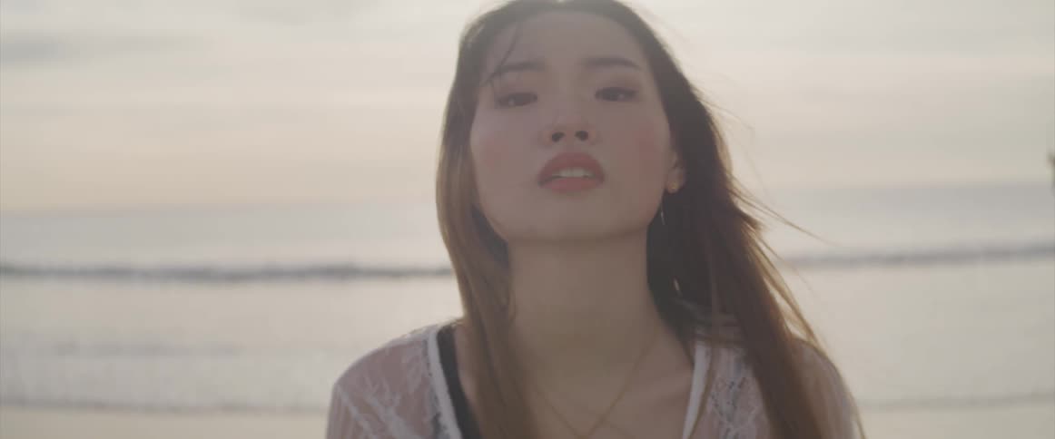 Asian woman with a black bikini and a white transparent dress faces the camera with eye contact on the beach