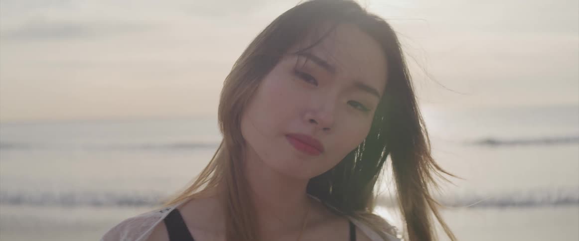 Asian woman with a black bikini and a white transparent dress standsand faces the camera smilingly on the beach