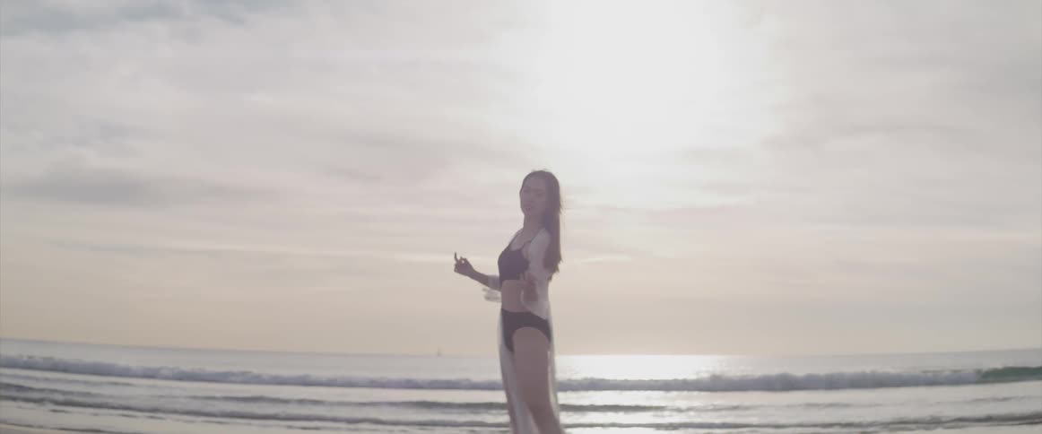 Asian woman with a black bikini and a white transparent dress dances and turns around on the beach at sunset