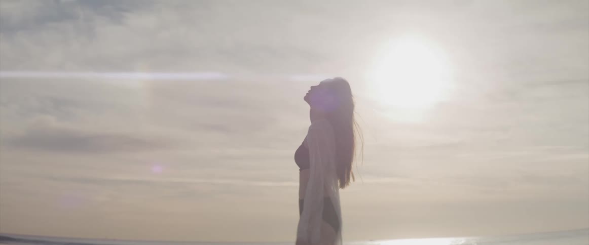 Woman in a bikini stands on the beach with her face tilted up towards the sky