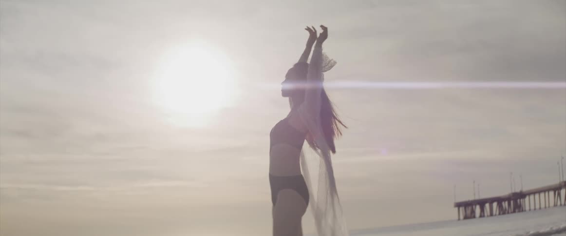 Woman wearing bikini an wrap stretches her arms out and up on beach while being backlit by glaring sun