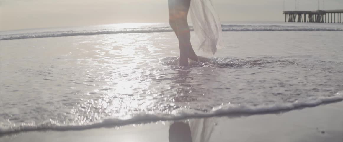 A woman in a swimsuit leisurely kicks the water as she walks through the tide on the beach on a cloudy day