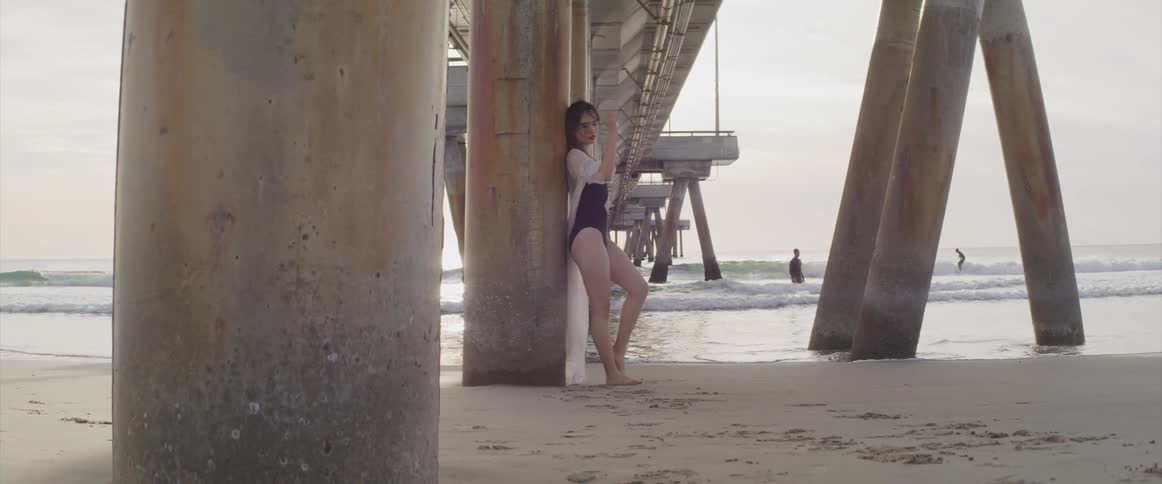 A young asian woman poses in a swimsuit beneath a large pier