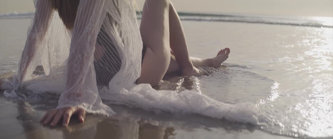 A woman in a swimsuit sits on the sand of the beach as the tide washes around her