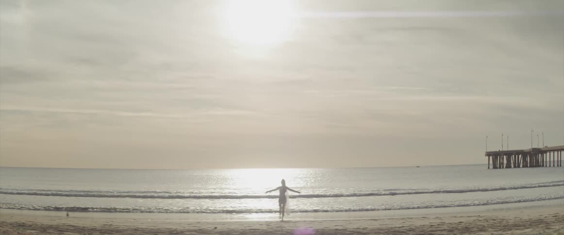 A woman in a swimsuit stands on the beach with her arms held wide before she slowly lowers them