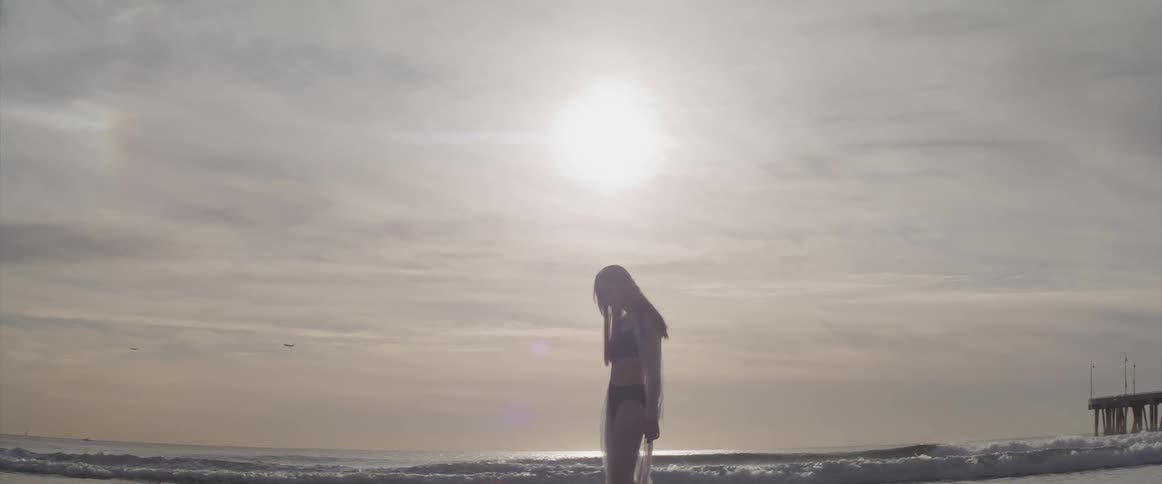 Asian woman with a black bikini and a white transparent dress standing on the beach is silhouetted against the cloudy sky