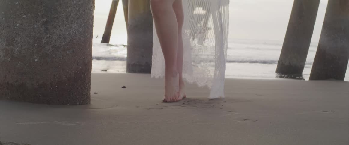 A woman poses on the beach under a pier while wearing a white lace coverup