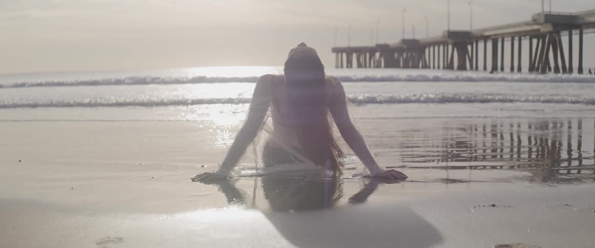Woman leans back on beach sunbathing as waves flow onto beach past pier