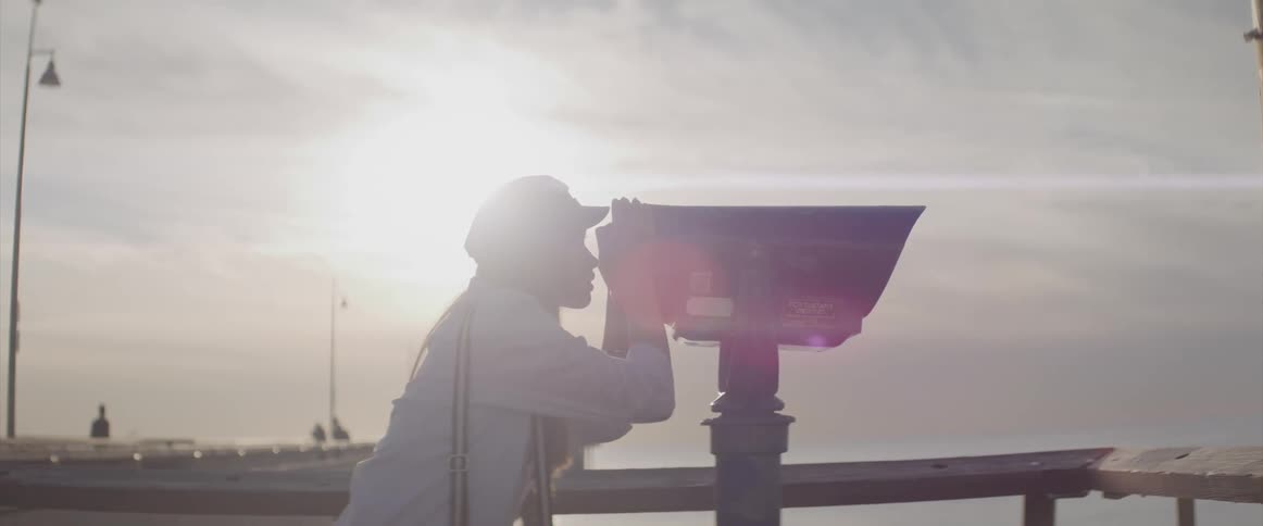 A woman in a boardwalk looking through binoculars with the sky and clouds in the background