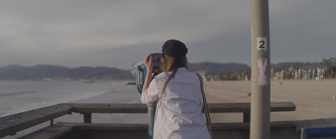 A woman standing on a dock on the beach looks through a telescope