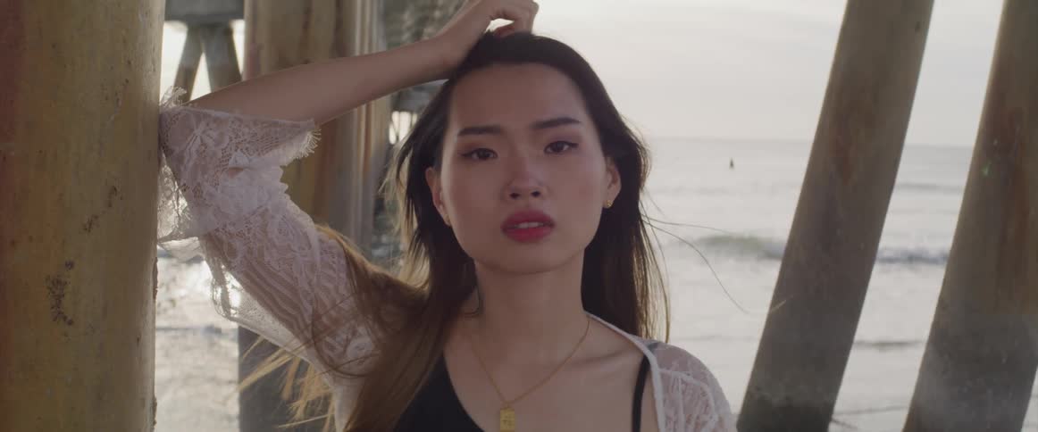 Asian woman with a black bikini and a white transparent dress faces the camera with eye contact under a pier on the beach