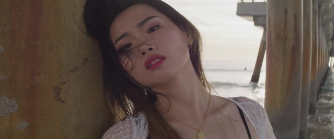 Asian woman with a black bikini and a white transparent dress faces the camera with eye contact under a pier on the beach