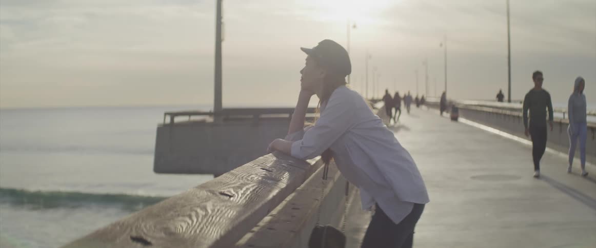 A young woman leans on a bridge and looks out at the water as other people walk along the bridge