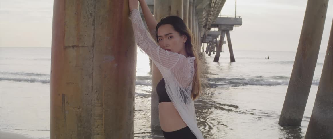 Woman wearing swimsuit and wrap leans against pier support while she poses