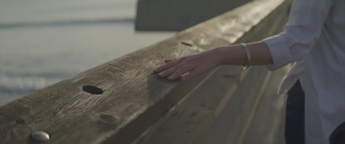 A woman runs her hand along the railing of a pier as she walks along and looks out at the water
