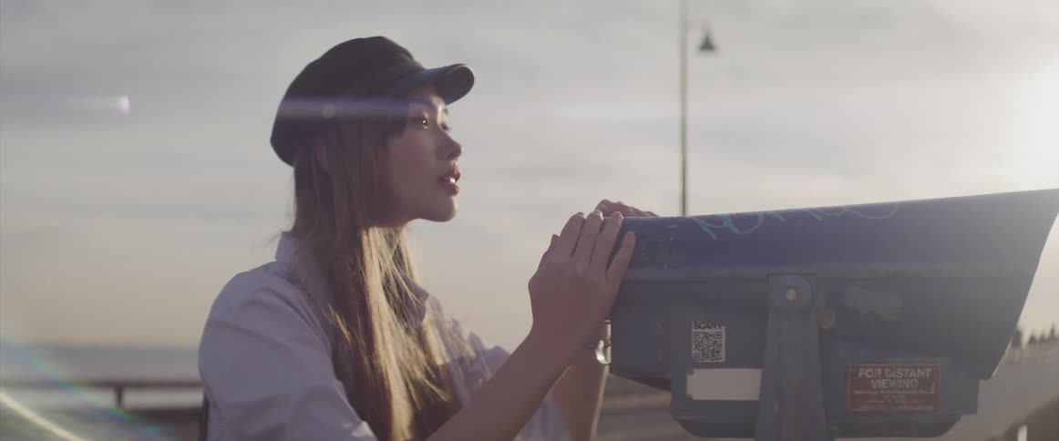 A woman with a hat standing in a boardwalk looking through binoculars with the sky in the background