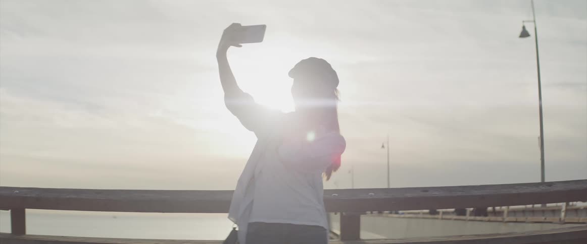 A woman stands on an oceanside pier holding up a cell phone to take a selfie