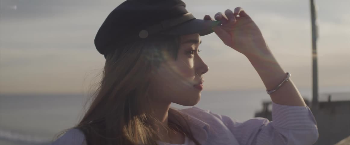 A young asian woman adjusts her student cap while leaning against a banister on the beach during the day
