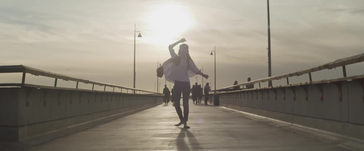 A teenage girl twirls around on a bridge with people walking and standing on it