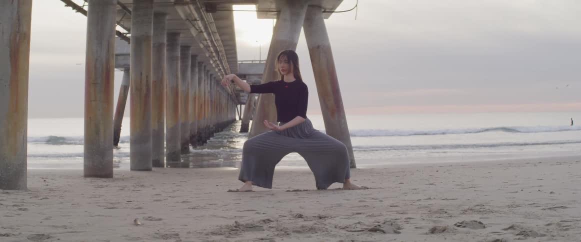 A woman performs meditation moves under a large ocean pier