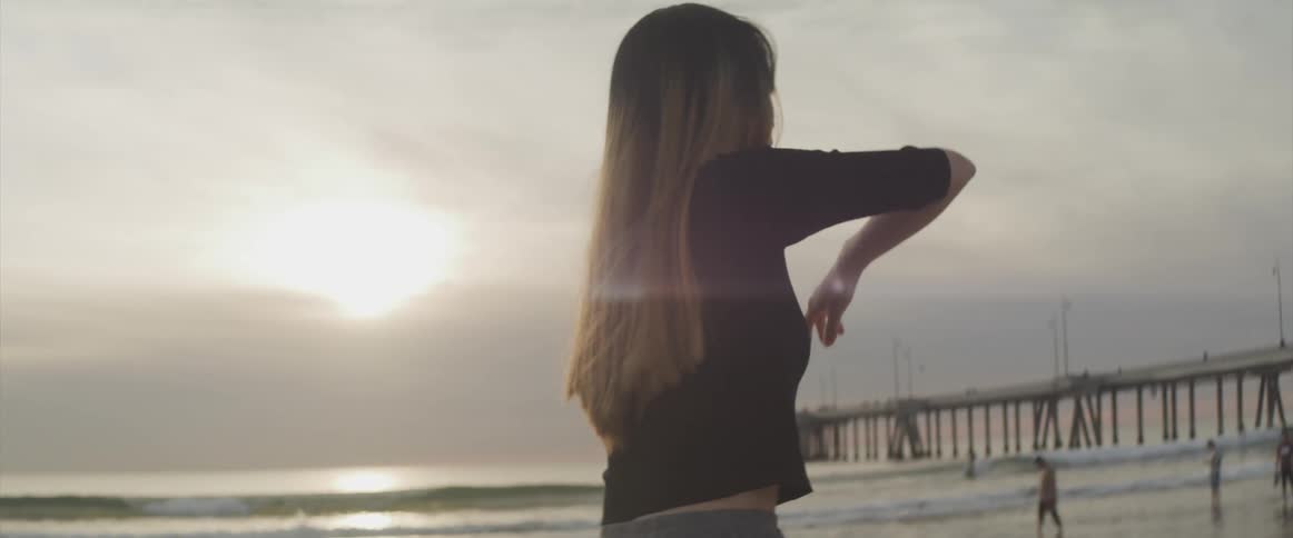 A woman practices tai chi on the beach while several people walk in the water