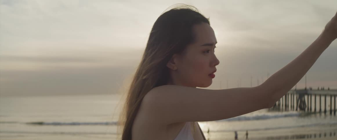 A young asian woman dances on the beach as the sun peaks through thick clouds in the sky behind her