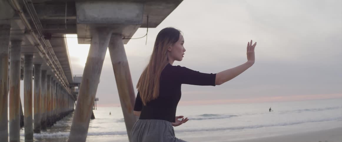 A woman does meditation moves standing alone on a beach under a pier