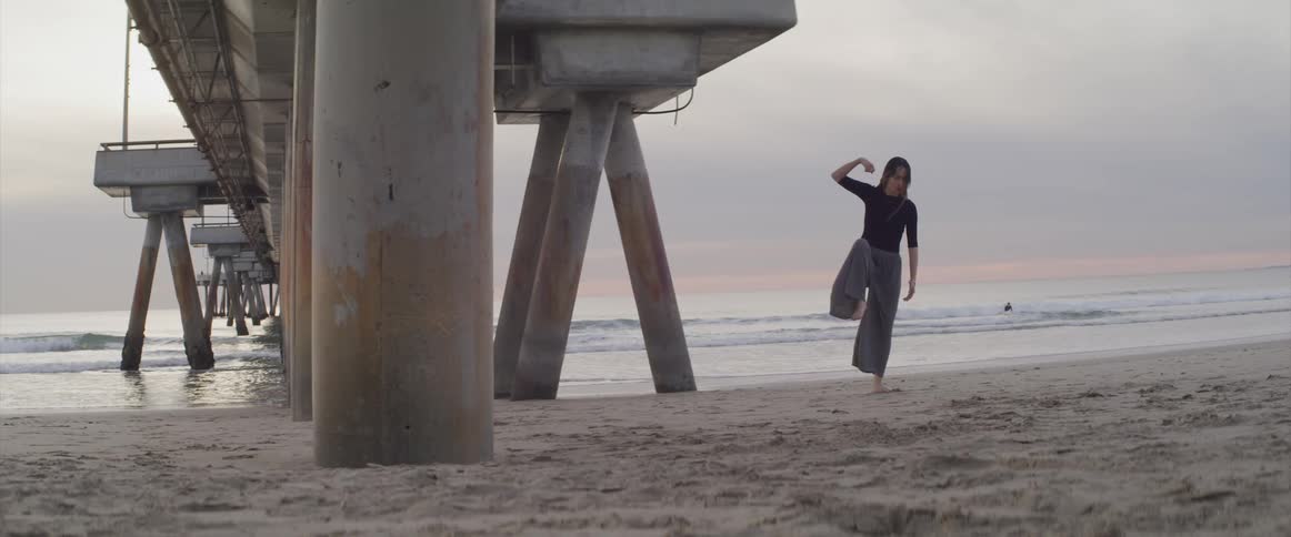 A woman does tai chi under a pier at the beach right next to the ocean