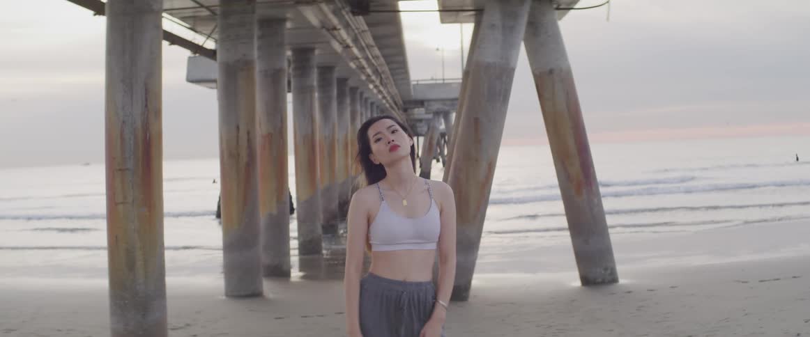 A young woman standing beneath a wooden pier poses on a sandy beach