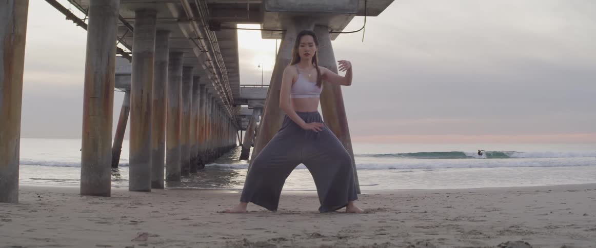 A young woman practices martial arts on the beach while another person surfs in the water
