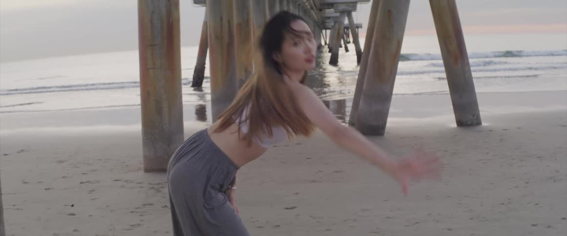 A young asian woman dances under a wooden pier on a beach during the day