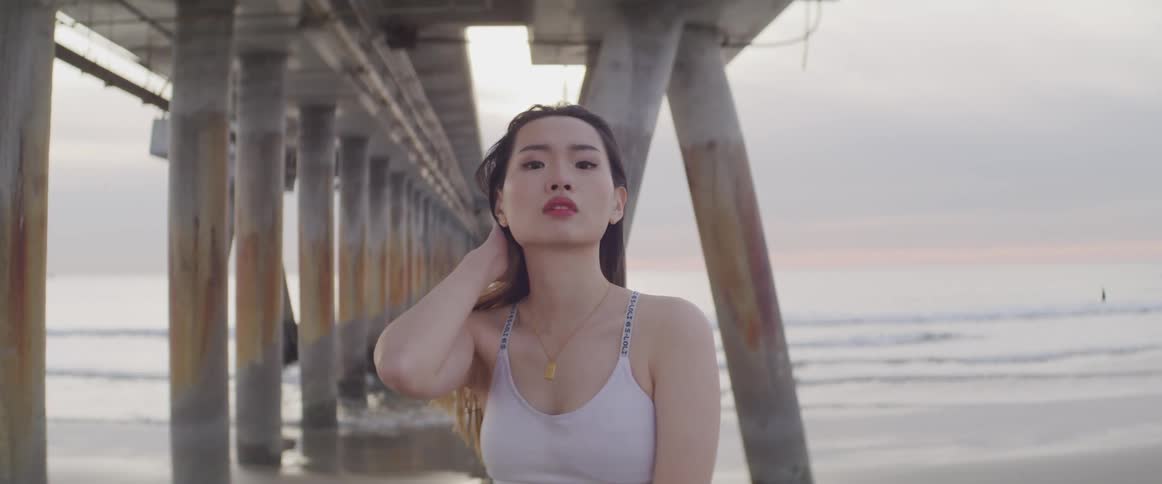 Asian woman with sports bra stands and throws hair back on the beach under a pier on a cloudy day