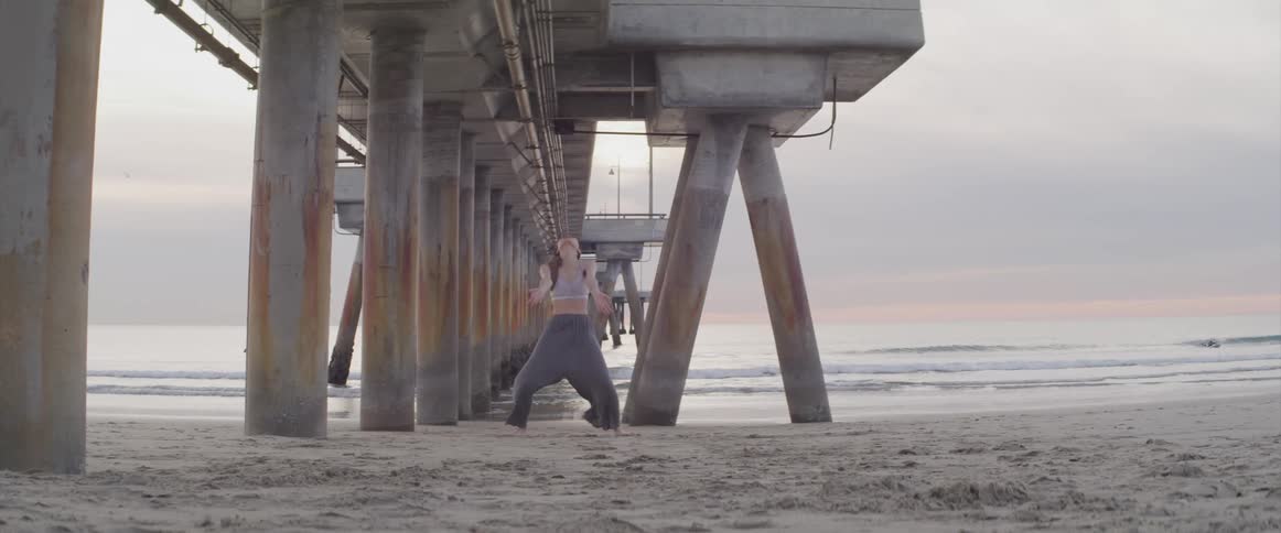 A teenage girl dancing on the beach as a person surfs in the water