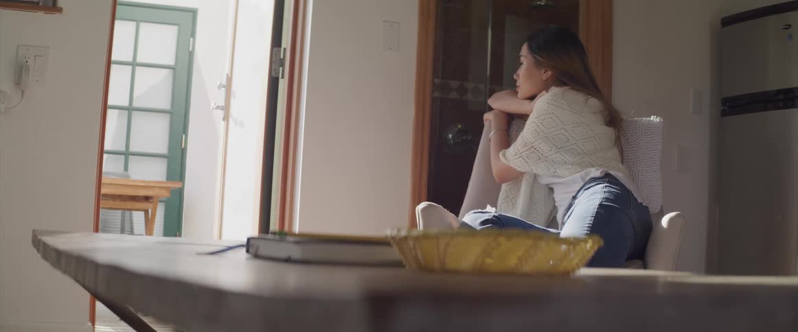 Asian woman with a white dress and jeans sits in an armchair and looks out the door