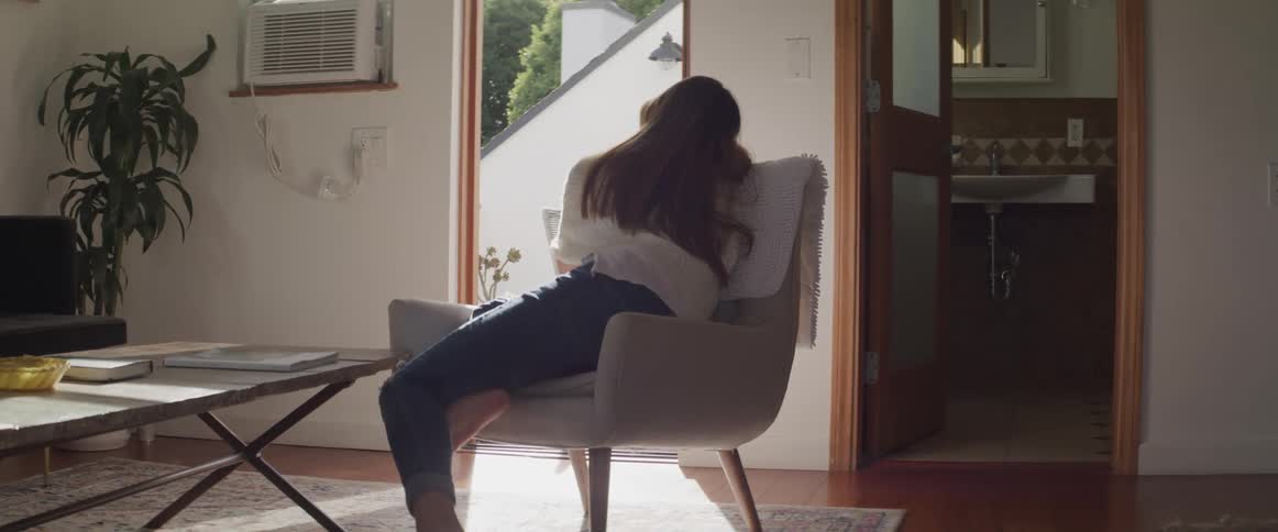 A young woman sits sideways in a gray upholstered chair in her living room and looks out the open front door
