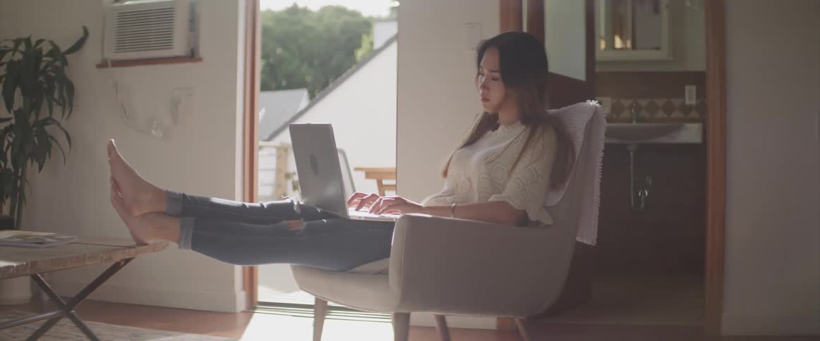 Depressed female sitting in chair in living room near the bathroom with her bare feet propped up on a table types on a laptop that is resting on top of her legs