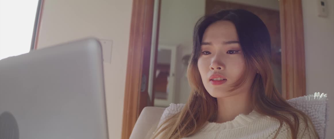 Asian woman with a white sweater sits in an armchair and uses a grey laptop