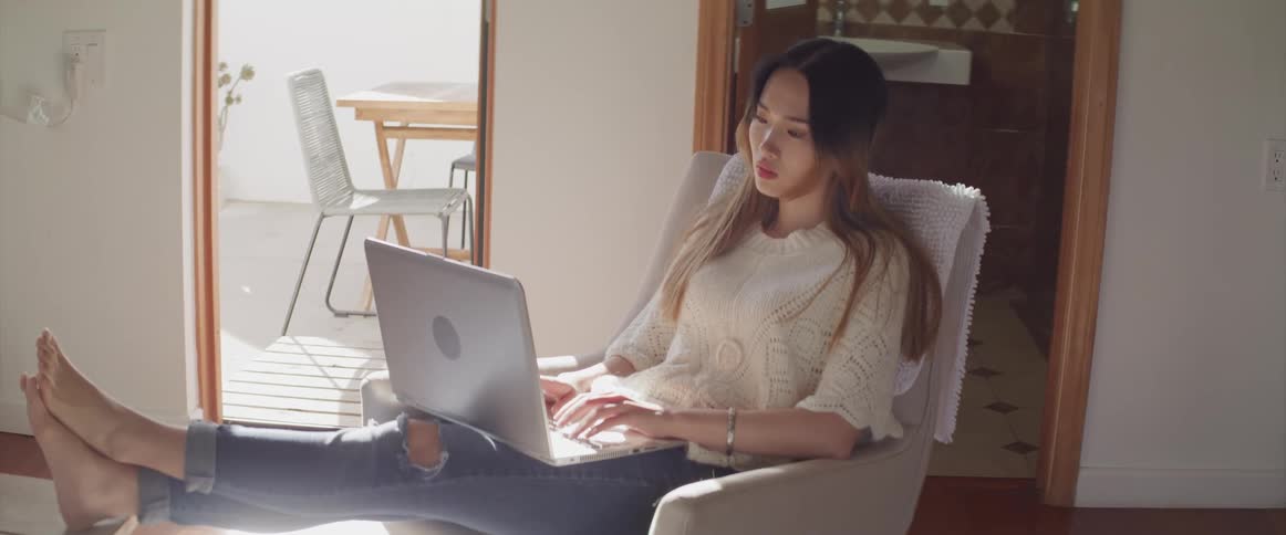 Woman sits with legs up typing on laptop held in her lap in home