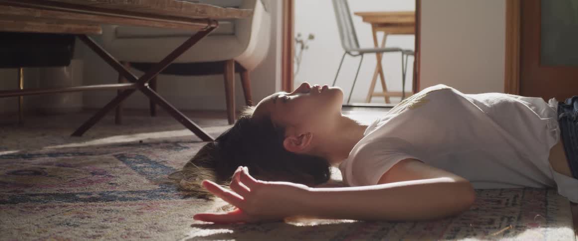 Asian woman with a white shirt and jeans lies on a carpet and looks at the ceiling in the living room