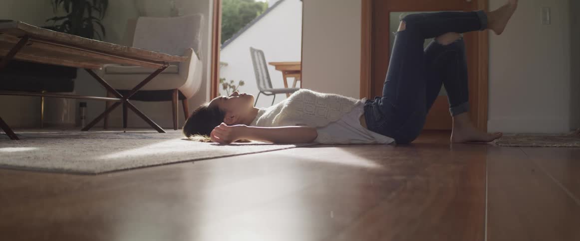 A young asian woman relaxes on her back on the floor of a living room on a sunny day