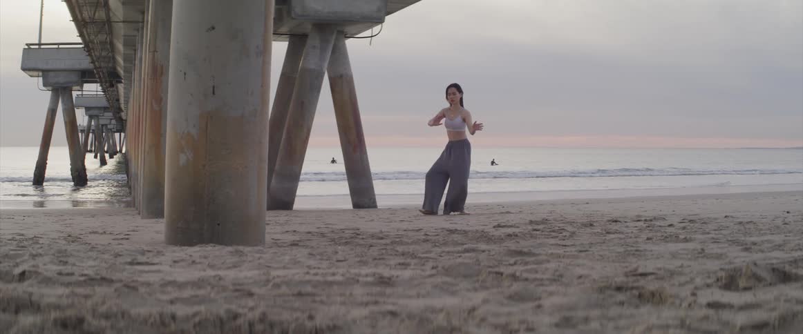Young woman practices tai chi on the sand beach at sunset under the pier next to the ocean
