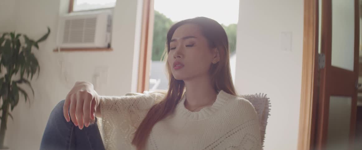 Asian woman with brown straight hair a white sweater and jeans sitting in an armchair looks at the camera