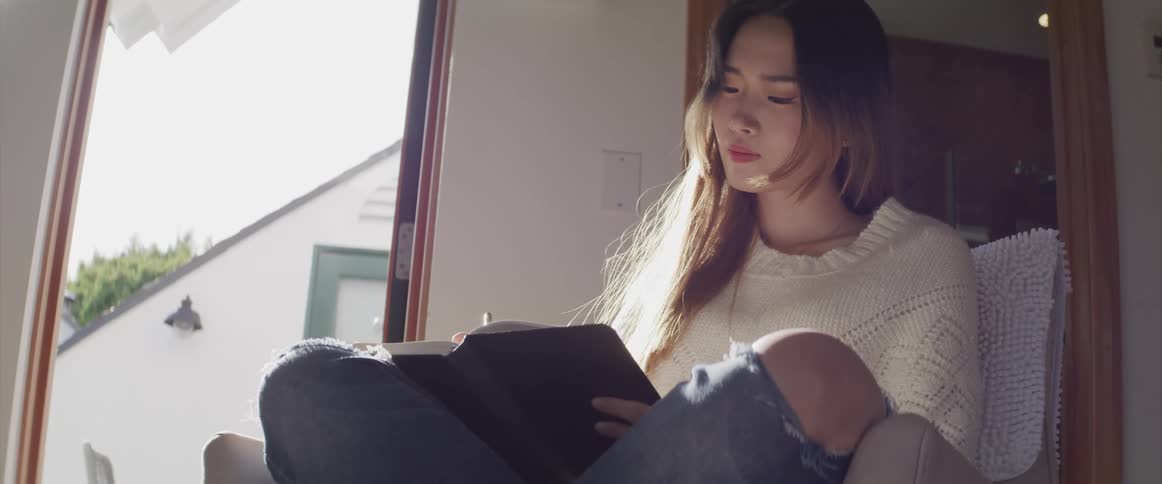 Asian girl with ripped jeans a white sweater sits in an armchair and reads a book in the living room