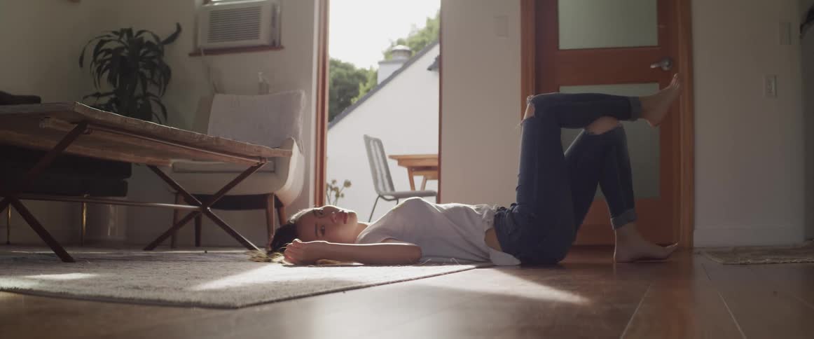 Asian woman with a white shirt and ripped jeans lies on a carpet and turns her head to the camera in the living room