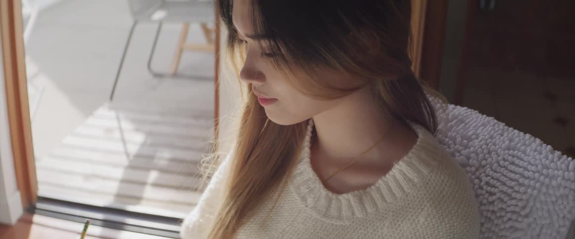 A young asian woman sits on a chair in a house while writing with a pencil
