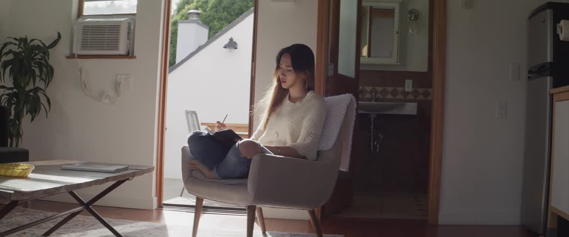 A young asian woman sits on a chair in the living room of a house while writing in a book
