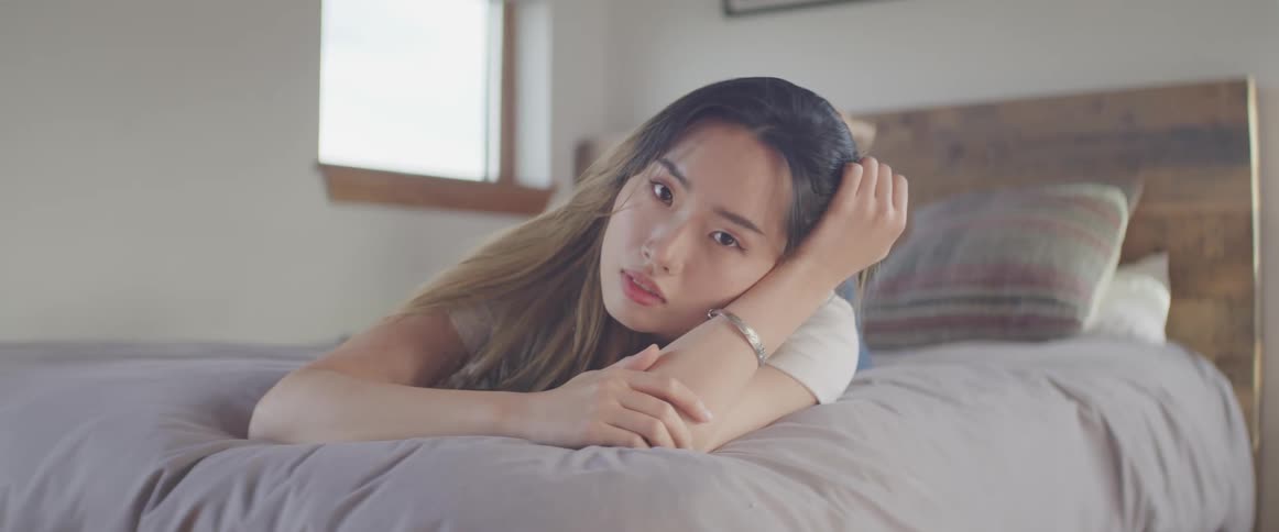 A young asian woman resting on her stomach on a bed raises her head to look into the camera