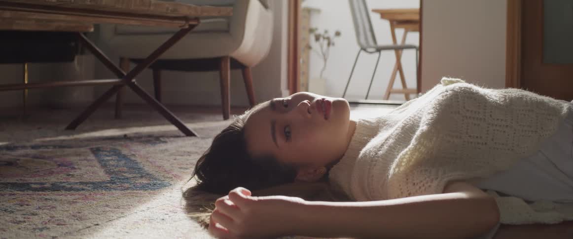 Asian woman with ripped jeans and a white sweatshirt lies on floor and looks at the camera in the living room