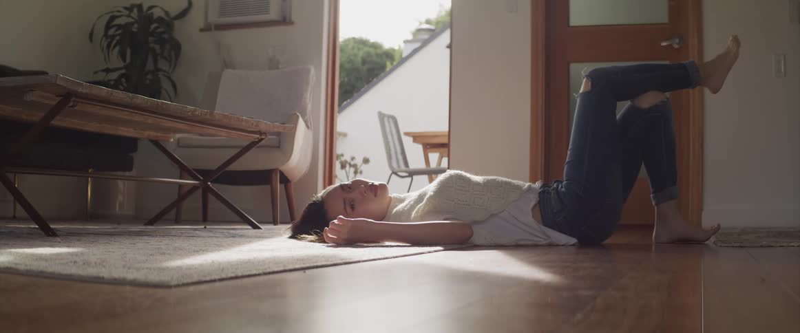 Asian woman with ripped jeans and a white sweatshirt lies on floor and looks at the camera in the living room