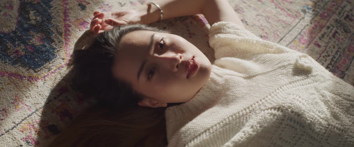 Sunlight from a window shines onto a young asian woman lying on a rug as she looks into the camera